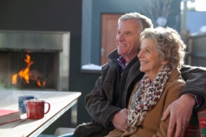 Two smiling people wearing light coats drink coffee sitting before an outdoor fireplace